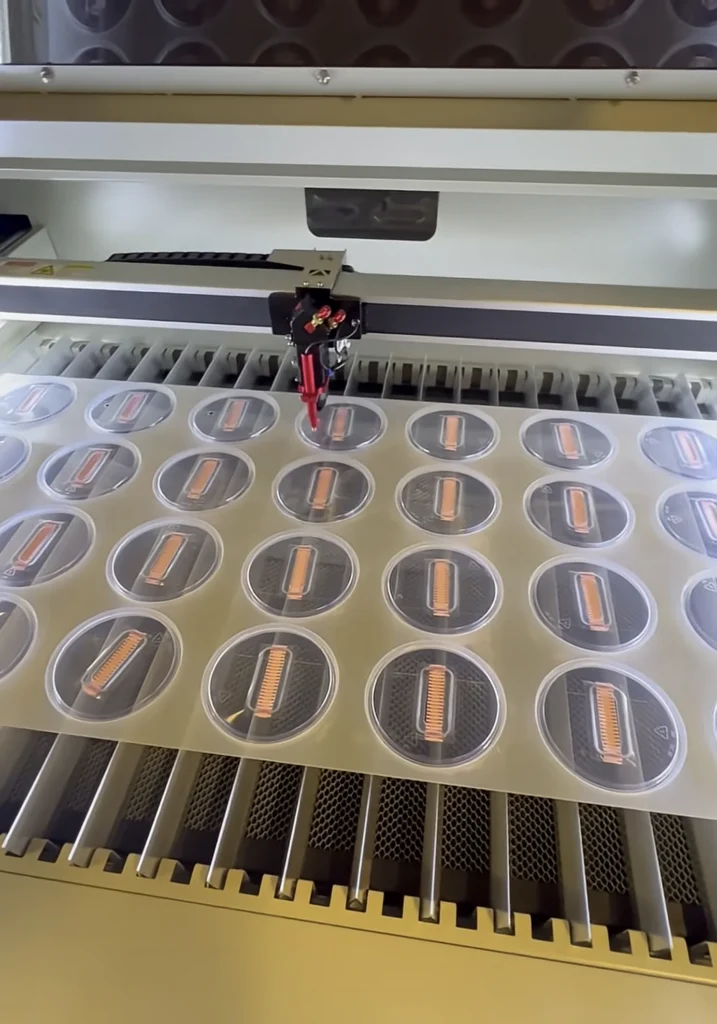 Our laser cutter cutting injection port holes in a sheet of grow kit tub lids with filter strips